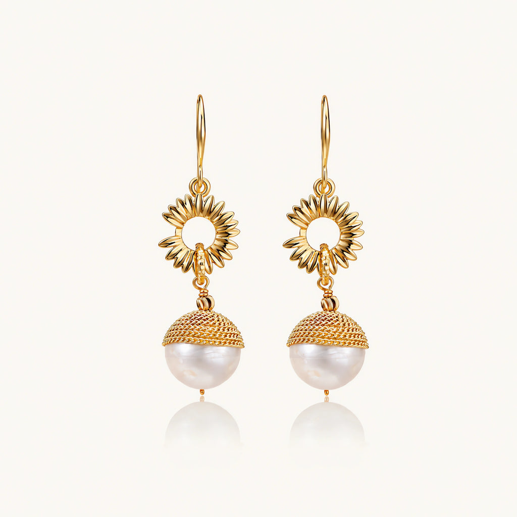 Gold sunburst charm earrings with baroque pearl drops on French hooks against cream background