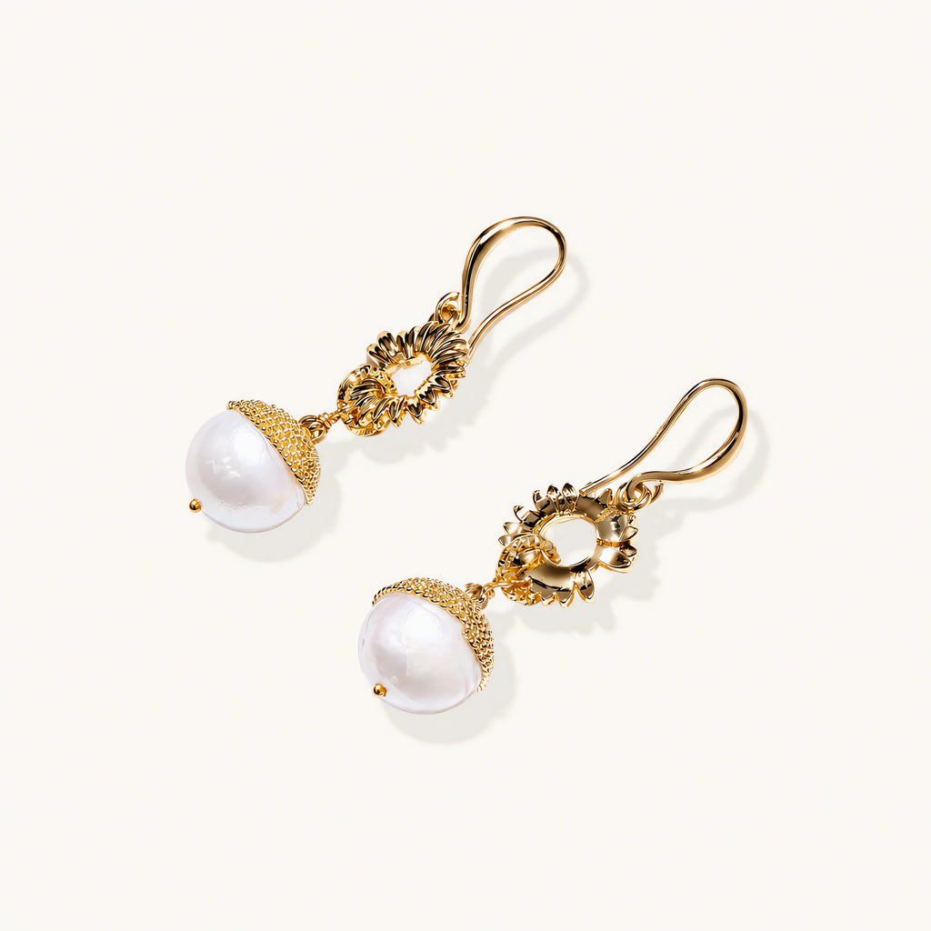 Gold sunburst charm earrings with baroque pearl drops on French hooks against cream background