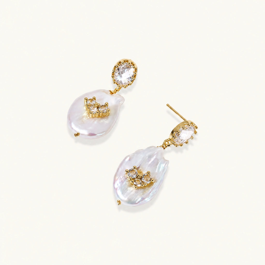 Gold-plated drop earrings with iridescent baroque pearls and crystal flower accents on cream background