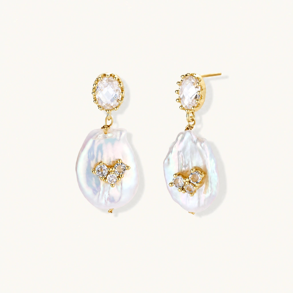 Gold-plated drop earrings with iridescent baroque pearls and crystal flower accents on cream background