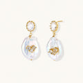 Gold-plated drop earrings with iridescent baroque pearls and crystal flower accents on cream background