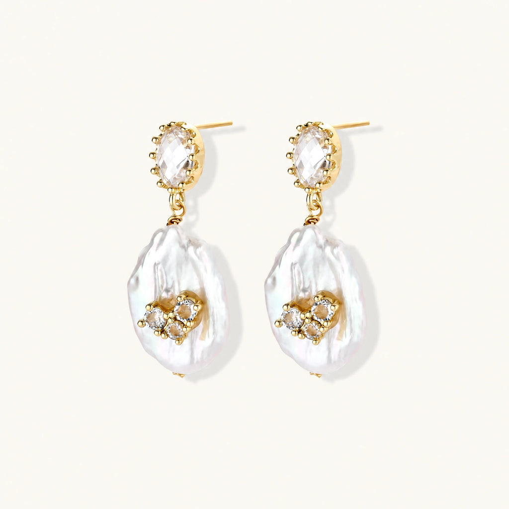Gold-plated drop earrings with iridescent baroque pearls and crystal flower accents on cream background