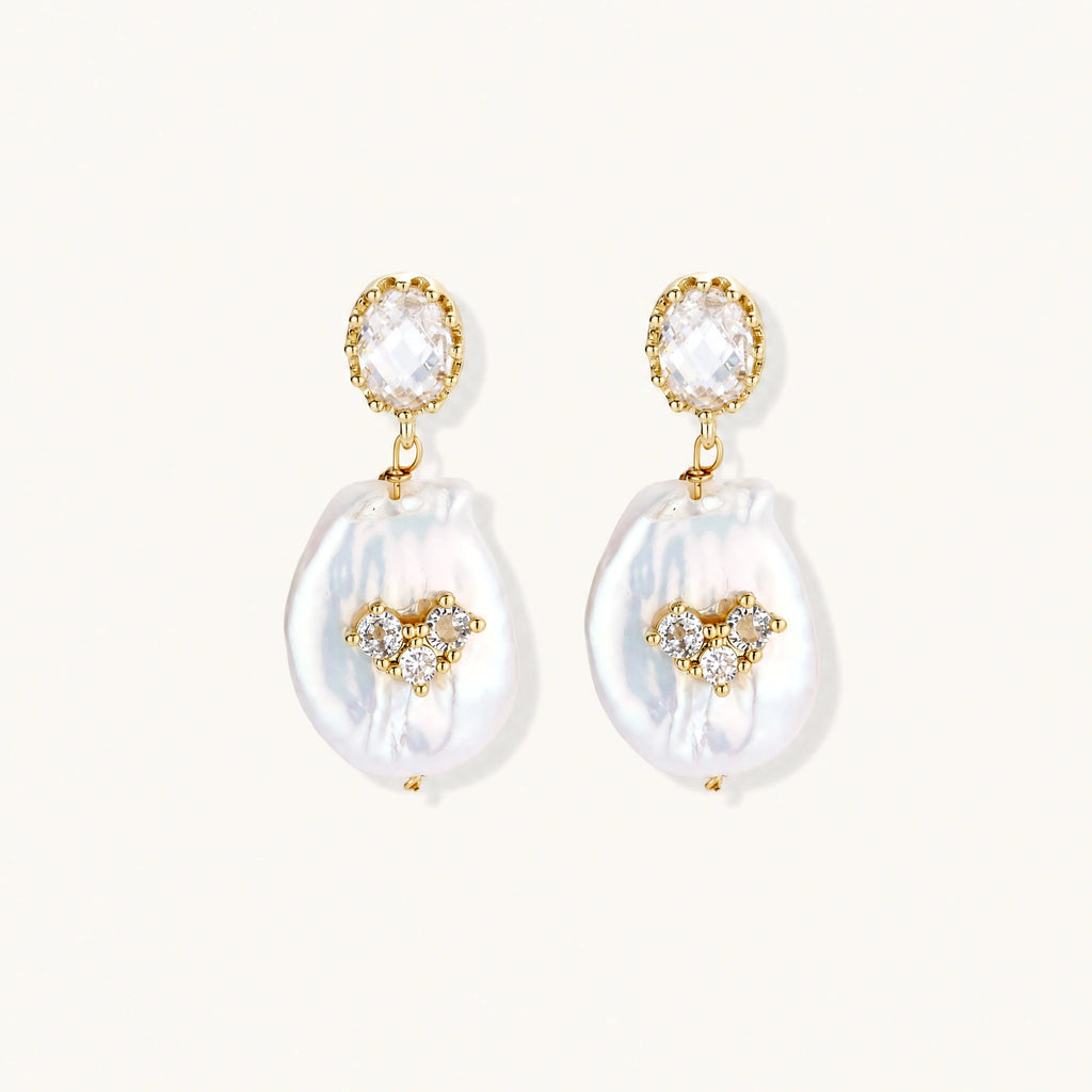 Gold-plated drop earrings with iridescent baroque pearls and crystal flower accents on cream background